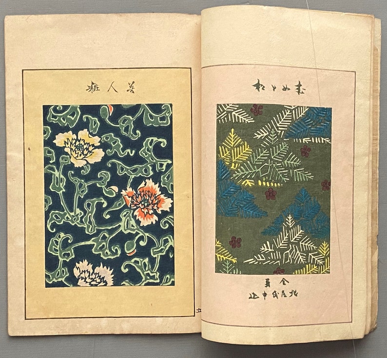 Op de afbeelding: Twee pagina's uit een vintage Japans boek met stoffenpatronen. De linkerpagina toont een bloemenpatroon met witte, gele, oranje en groene bloemen op een donkerblauwe achtergrond. De rechterpagina toont een groene achtergrond met een patroon van gestileerde dennennaalden in tinten groen, geel en blauw.