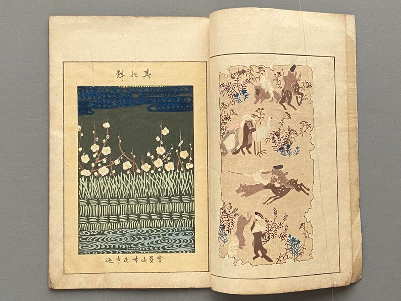 Op de afbeelding: Een open boek met twee pagina's die Japanse illustraties tonen. De linkerpagina toont een landschap met witte bloemen en een blauwe lucht. De rechterpagina toont een jachtsc&egrave;ne met mensen te paard die herten achtervolgen.