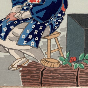 May include: A Japanese woodblock print depicting a person wearing a blue and white patterned robe sitting on a wooden stool. The person is holding a fan in their hand. The print is signed in Japanese characters.