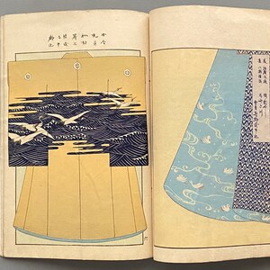 Op de afbeelding: Twee pagina's uit een Japans boek met verschillende ontwerpen voor een kimono. De linkerpagina toont een kimono met een patroon van witte vogels die over blauwe golven vliegen. De rechterpagina toont een close-up van de onderkant van een kimono met een vergelijkbaar patroon van witte vogels en blauwe golven.