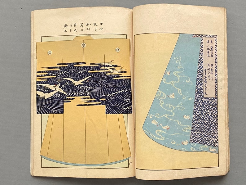 Op de afbeelding: Twee pagina's uit een Japans boek met verschillende ontwerpen voor een kimono. De linkerpagina toont een kimono met een patroon van witte vogels die over blauwe golven vliegen. De rechterpagina toont een close-up van de onderkant van een kimono met een vergelijkbaar patroon van witte vogels en blauwe golven.