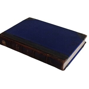 Puede incluir: Un libro antiguo de tapa dura con una cubierta azul oscuro y letras doradas en el lomo. El libro est&aacute; encuadernado en cuero marr&oacute;n y tiene un aspecto desgastado y envejecido.