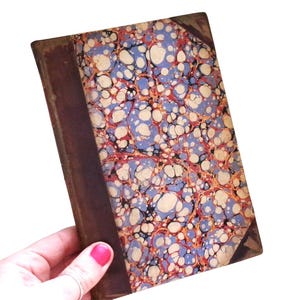 May include: Antique book with a marbled cover featuring a pattern of blue, white, and red swirls. The spine and corners are a dark brown leather. The book is held in a hand.