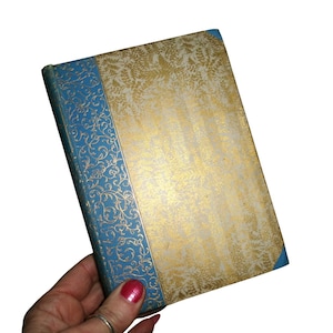 May include: A vintage book with a gold and white cover featuring a floral pattern. The spine and corners are blue with gold floral detailing. The book is held in a hand.