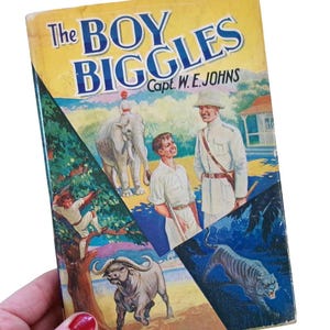May include: A vintage copy of "The Boy Biggles" by Capt. W.E. Johns. The book cover displays illustrations of an elephant, a man in uniform, a boy, a tiger, and a water buffalo. The cover is yellow and blue, with signs of wear and tear.