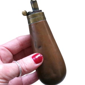 Könnte beinhalten: Ein antikes Pulverfläschchen aus Kupfer mit einem konisch zulaufenden, abgerundeten Körper und einem Messingmechanismus an der Oberseite. Das Metallteil oben verfügt über einen kleinen Ausguss und einen Hebel zur Dosierung mit Patina.