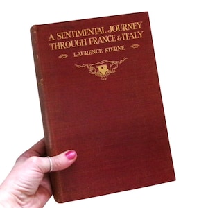 Pode incluir: Um livro de cor castanho-avermelhado intitulado "A Sentimental Journey Through France & Italy" de Laurence Sterne. A capa apresenta letras douradas e um emblema decorativo. O livro é segurado em uma mão.