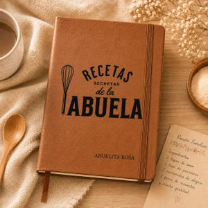 May include: A tan leather-bound notebook with a vertical elastic closure. The cover features a black whisk icon and the text "RECETAS SECRETAS de la ABUELA" with the name "ABUELITA ROSA" printed below in a clean, dark sans-serif font.