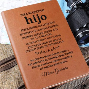 May include: A brown leather journal with the words "PARA MI QUERIDO hijo" in black. The journal has a textured surface and contains a heartfelt message. A camera and map are visible in the background.