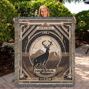 May include: Woven blanket featuring a deer silhouette in a mountain landscape, with the words "ACILIE" and "CHIIE" at the top, and "HUEMUL CHILENO" at the bottom. The blanket has a fringed edge and a geometric design.