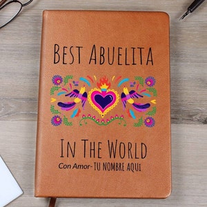 Puede incluir: Un cuaderno de piel sint&eacute;tica color canela con el texto negro BEST ABUELITA IN THE WORLD y Con Amor- TU NOMBRE AQUI. La portada muestra un gr&aacute;fico colorido de un coraz&oacute;n central con llamas flanqueado por dos p&aacute;jaros y flores en rosa, azul y amarillo.