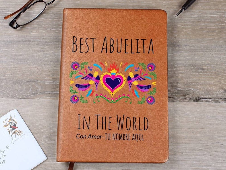 Puede incluir: Un cuaderno de piel sint&eacute;tica color canela con el texto negro BEST ABUELITA IN THE WORLD y Con Amor- TU NOMBRE AQUI. La portada muestra un gr&aacute;fico colorido de un coraz&oacute;n central con llamas flanqueado por dos p&aacute;jaros y flores en rosa, azul y amarillo.