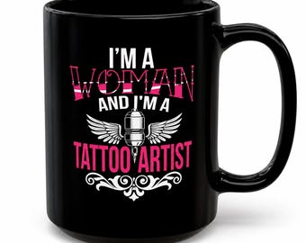 May include: A black ceramic mug featuring the white and pink text I'M A WOMAN AND I'M A TATTOO ARTIST. The design includes a central graphic of a tattoo machine with wings and a decorative white flourish at the bottom of the mug's front surface.
