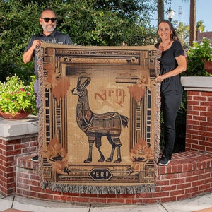 May include: A tan and brown woven throw blanket featuring a stylized llama design. The blanket has a rectangular shape with a decorative border and the word "PERU" at the bottom. The blanket is being held up by two people.