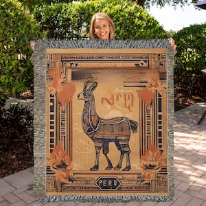 May include: A woven throw blanket with a vintage-style illustration of a llama in front of a mountain range. The llama is brown and black with geometric patterns. The blanket has a brown, black, and white border with a geometric pattern. The text "PERU" is in a hexagon shape at the bottom of the blanket.