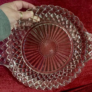 May include: A clear, vintage glass serving dish with a diamond pattern around the rim and a sunburst design in the center. The dish has two handles and is held by a hand. The background is a deep red.