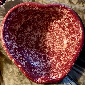 以下が含まれることがあります： ハート型の陶器のボウル。ボウルは、白い斑点のある濃いバーガンディ色です。ボウルの縁は滑らかで丸みを帯びています。ボウルは、サービングまたはディスプレイに使用される可能性があります。