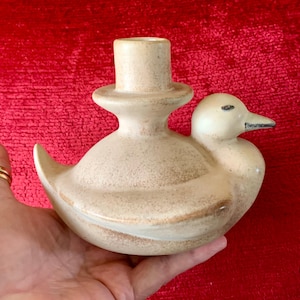 May include: A ceramic duck-shaped candle holder in a speckled beige colour. The duck has a cylindrical candle holder on its back and a black beak. The candle holder is held in a hand against a red background.