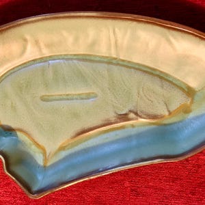 Puede incluir: Una fuente de cerámica con una forma asimétrica única. La fuente presenta un esmalte de dos tonos, con una sección superior de color amarillo claro y una sección inferior azul-verde. El borde es de color marrón oscuro. La fuente tiene una hendidura rectangular.