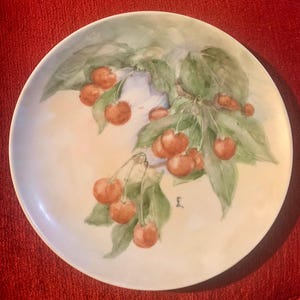 Könnte beinhalten: Ein weißer Keramikteller mit einem handgemalten Design aus Kirschen und grünen Blättern. Die Kirschen sind leuchtend rot und die Blätter sind in verschiedenen Grüntönen gehalten. Der Teller hat einen leicht gewellten Rand.