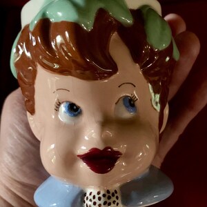 May include: A ceramic figural mug shaped like a head with brown hair, light green leaves on top, blue eyes, red lips, and a blue collar with a small white and black polka dot detail. The glossy finish shows fine surface cracks throughout the glaze.