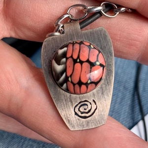 May include: A silver pendant with a black leather cord. The pendant features a round, colourful glass inlay with a swirl design. The pendant has a spiral design etched on the back.
