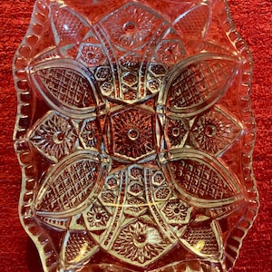 May include: A clear glass dish with a scalloped edge and intricate cut-glass design. The dish has a geometric pattern with a central starburst.