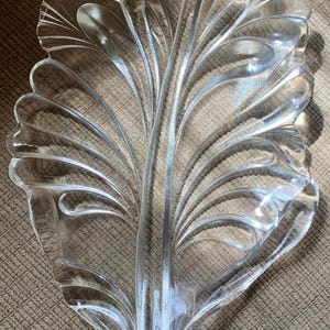 Op de afbeelding: Decoratieve serveerschaal van helder glas met een bladachtig ontwerp. De schaal heeft een centrale steel met gebogen, waaiervormige secties die naar buiten uitsteken. Het glas is transparant en ligt op een gestructureerd oppervlak.
