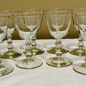May include: A set of ten clear glass wine goblets. Each goblet has a round base, a stem with a series of spherical accents, and a flared bowl. The goblets are arranged on a white surface, suggesting a formal dining setting.