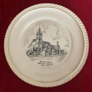 Pode incluir: Um prato de cerâmica branco com uma borda dourada e uma ilustração em preto e branco de uma igreja. O texto "Methodist Church, Monroeville, Indiana 1847 - 1957" está impresso no prato.