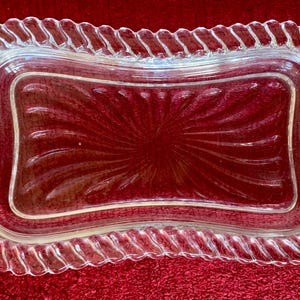 May include: A clear glass butter dish with a decorative rope-like border. The dish has a swirl pattern on the inside.
