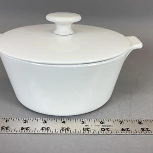 May include: A white ceramic casserole dish with a lid. The dish has a rounded shape with two small handles and a knob on the lid. A ruler is placed next to the dish, showing measurements in centimetres.