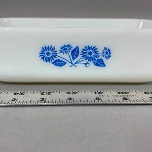 Pode incluir: Assadeira retangular branca com design floral azul. A assadeira tem uma alça em cada lado. Uma régua é colocada na frente da assadeira. A assadeira tem aproximadamente 25 cm de comprimento.