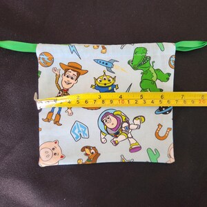 May include: A light blue drawstring bag featuring Toy Story characters. The bag is approximately 9 inches wide. The bag has a green drawstring closure.