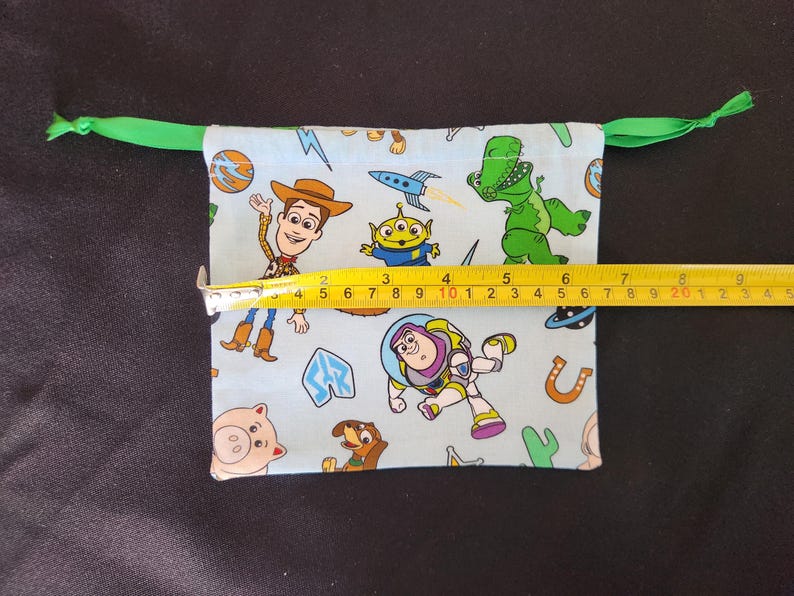 May include: A light blue drawstring bag featuring Toy Story characters. The bag is approximately 9 inches wide. The bag has a green drawstring closure.