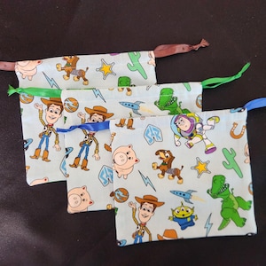 May include: Three light blue drawstring bags featuring characters from the Toy Story movie. The bags have brown, green, and blue ribbons. The bags are decorated with images of Woody, Buzz Lightyear, Rex, and other characters.