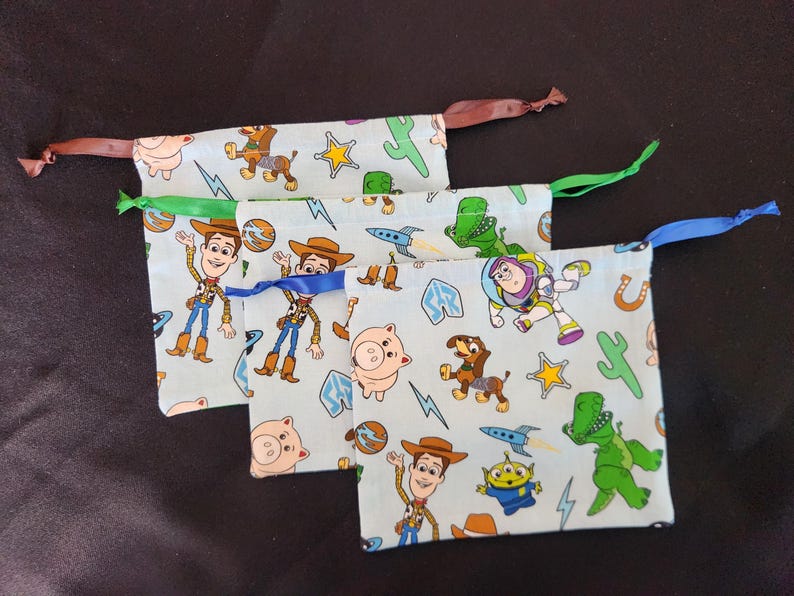 May include: Three light blue drawstring bags featuring characters from the Toy Story movie. The bags have brown, green, and blue ribbons. The bags are decorated with images of Woody, Buzz Lightyear, Rex, and other characters.