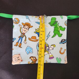 May include: A light blue drawstring bag featuring Toy Story characters like Woody, Buzz Lightyear, and Rex. The bag is approximately 4 inches tall and has a green drawstring closure.