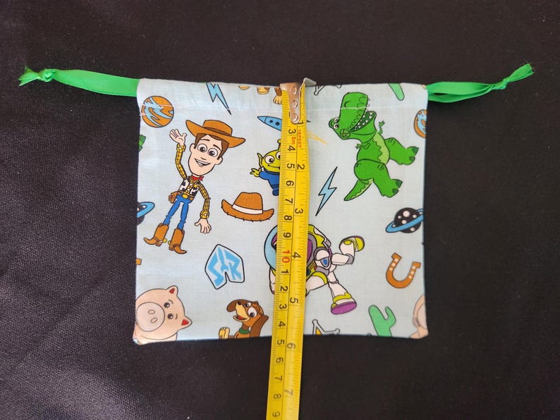 May include: A light blue drawstring bag featuring Toy Story characters like Woody, Buzz Lightyear, and Rex. The bag is approximately 4 inches tall and has a green drawstring closure.