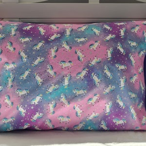 May include: A pink and purple pillowcase with a white unicorn pattern and a navy blue border. The unicorns are surrounded by stars and a watercolor-like background.