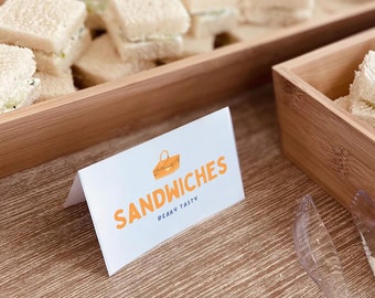May include: A wooden serving tray filled with small sandwiches, alongside a white card with the word "SANDWICHES" in orange lettering. The card also features a picnic basket illustration and the words "BEARY TASTY".
