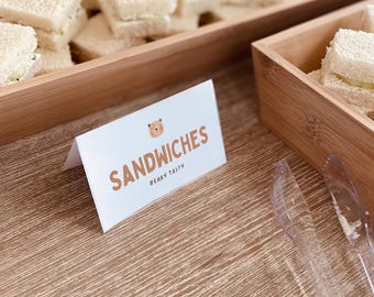 May include: A wooden serving tray filled with small sandwiches, alongside a sign that reads "SANDWICHES" with a bear illustration. The sign also includes the text "BEARY TASTY". Clear plastic serving utensils are visible.