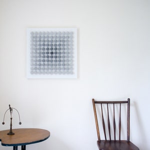 May include: A white canvas with a geometric pattern of gray circles in a grid formation. The canvas is hanging on a white wall. A wooden chair with a slatted back and a wooden table with a metal base are in the foreground.