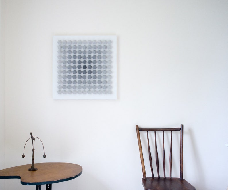 May include: A white canvas with a geometric pattern of gray circles in a grid formation. The canvas is hanging on a white wall. A wooden chair with a slatted back and a wooden table with a metal base are in the foreground.