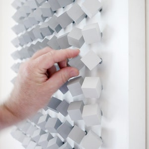 May include: A hand reaches out to touch a wall art piece made of small, gray, wooden cubes. The cubes are arranged in a staggered pattern, creating a textured and modern look.