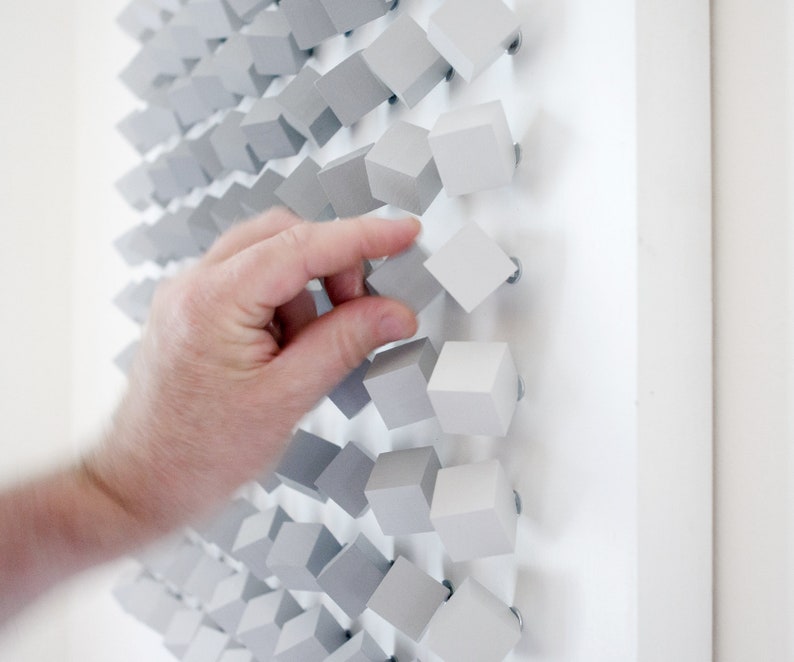 May include: A hand reaches out to touch a wall art piece made of small, gray, wooden cubes. The cubes are arranged in a staggered pattern, creating a textured and modern look.