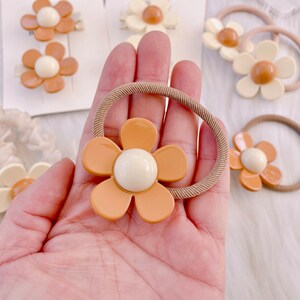 May include: A collection of hair accessories featuring daisy-shaped embellishments. The accessories include hair ties and barrettes in shades of orange, cream, and tan. The daisy centers are a creamy white color.