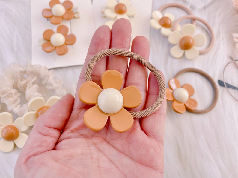 May include: A collection of hair accessories featuring daisy-shaped embellishments. The accessories include hair ties and barrettes in shades of orange, cream, and tan. The daisy centers are a creamy white color.