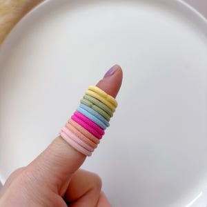 K&ouml;nnte beinhalten: Ein Stapel bunter Haargummis in Pastellfarben wie Rosa, Gelb, Blau und Gr&uuml;n. Die Haargummis sind aus weichem, dehnbarem Material und perfekt zum Festhalten von Haaren.
