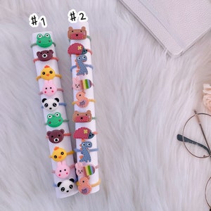 May include: Two white cylindrical displays labeled #1 and #2, each holding a vertical stack of colorful elastic hair ties. The hair ties feature various small, flat, cartoon-style animal and creature charms, including frogs, bears, chicks, bunnies, pandas, dinosaurs, and hedgehogs.